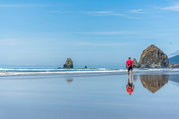 Man in a red T-shirt is walking along the water on the Northwest coast of the Pacific Ocean with rocks sticking out of the water reflected in the mirror water