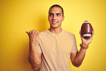 Poster - Young rugby player man holding a football ball over yellow isolated background pointing and showing with thumb up to the side with happy face smiling