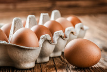 Brown Eggs In Carton Box. Broken Egg With Yolk At Background.