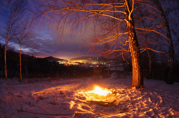 Wall Mural - Night fire on the mountain on the background of the bright sunset town village under a dark sky