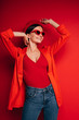 © Anton - Vertical picture of happy attractive cheerful young woman in red clothes and sunglasses posing on camera. Look to right and simle. Hands up. Isolated over red background.