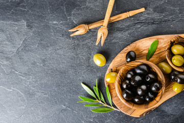 Wall Mural - Fresh green and black olives on dark stone table or board. Olive leaves, wooden pickers on black blackground.