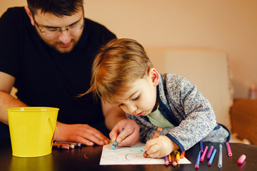 Wall Mural - Small boy and his uncle or father sitting by the table at home playing with crayons color pencils drawing and learning family activities having fun