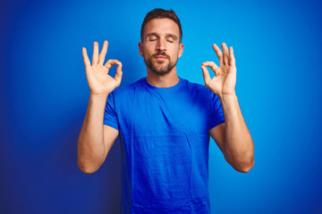 Poster - Young handsome man wearing casual t-shirt over blue isolated background relax and smiling with eyes closed doing meditation gesture with fingers. Yoga concept.