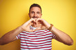 © Krakenimages.com - Young handsome man wearing casual red striped t-shirt over yellow isolated background smiling in love doing heart symbol shape with hands. Romantic concept.
