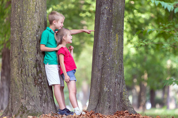 Portrait of two pretty cute children boy and girl standing near big tree trunk in summer park outdoors.