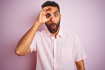 Wall Mural - Young indian man wearing casual shirt standing over isolated pink background doing ok gesture shocked with surprised face, eye looking through fingers. Unbelieving expression.