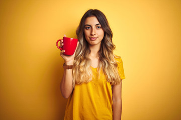 Sticker - Young beautiful woman holding  red cup of coffee over yellow isolated background with a confident expression on smart face thinking serious