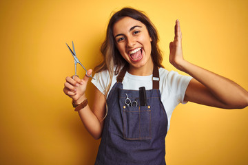 Poster - Young beautiful hairdresser woman holding scissors over yellow isolated background very happy and excited, winner expression celebrating victory screaming with big smile and raised hands