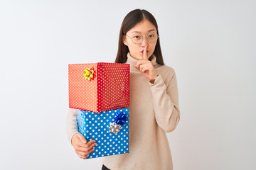 Canvas Print - Young chinese woman holding birthday gifts over isolated white background asking to be quiet with finger on lips. Silence and secret concept.