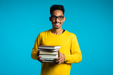 African student on blue background in the studio holds stack of university books from library. Guy smiles, he is happy to graduate.