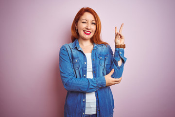 Sticker - Youg beautiful redhead woman wearing denim shirt standing over isolated pink background smiling with happy face winking at the camera doing victory sign. Number two.