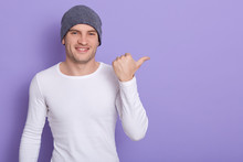 Portrait Of Stylish Handsome Young Man Osing Isolated Over Lilac Background. Male Smiling And Points Aside, Wears White Casual Long Sleeve Shirt And Gray Cap. Copy Space For Advertisment Or Promotion.