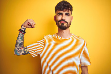 Sticker - Young man with tattoo wearing striped t-shirt standing over isolated yellow background Strong person showing arm muscle, confident and proud of power