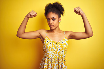 Sticker - African american woman wearing casual floral dress standing over isolated yellow background showing arms muscles smiling proud. Fitness concept.