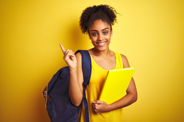 Wall Mural - American student woman wearing backpack holding notebook over isolated yellow background very happy pointing with hand and finger to the side
