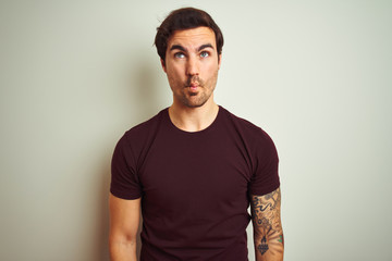 Wall Mural - Young handsome man with tattoo wearing purple casual t-shirt over isolated white background making fish face with lips, crazy and comical gesture. Funny expression.