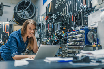 Portrait of beautiful working woman in the bicycle store. Young female mechanic with laptop. Female Bicycle Mechanic. Craftswoman working using a laptop in the garage