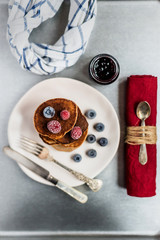 Wall Mural - Delicious buckwheat pancakes with frozen raspberries and blueberries on a plate, red napkin, fork, knife, spoon, a small jar of jam and a towel. Top view high key photo on grey background