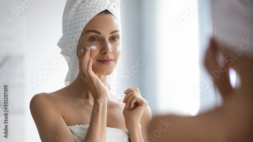Fotografía Attractive young adult woman applying facial cream looking in mirror