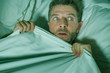  stressed and scared man alone in bed awake at night in fear after having a nightmare feeling paranoid holding the blanket in funny panic face expression