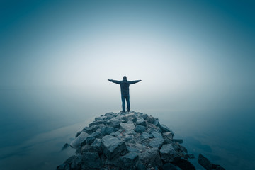 Man, standing on the stone heap at the foggy river bank. Long exposure shot.