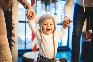 Wall Mural - Funny little baby boy 1 year old learning walk home in winter in a decorated New Year house. Young family dad and mom hold by the hands of his son in the loft interior wooden floor near the window