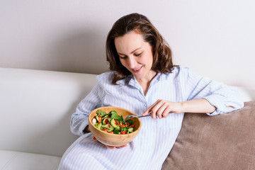 pregnant smiling woman sitting on sofa eating healthy vegetable salad in wooden bowl