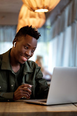 Wall Mural - Cheerful young african american student man in headphones using computer in cafe. Online video chat with friends