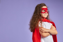 Portrait Of Beautiful Kid With Curly Hear Wearing Red Cloak And Red Mask Looking Side, Purple Background. Children Interested In Cosmos