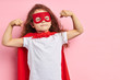Leinwandbild Motiv Beautiful little curly girl wearing red hero suit and mask showing how she is strong isolated over pink background