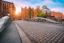 Arch Bridge Over Canals With Cobbled Road In The Speicherstadt Of Hamburg, Germany, Europe. Historical Red Brick Building Lit By Golden Sunset Light Near Water Castle Palace
