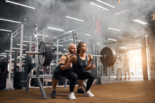 Rude Brutal Trainer With Bald Head And Thick Brunette Beard Standing Behind Young Female Powerlifter, Backing Her Up, Controlling Process Of Squatting Using Heavy Barbell In Gym, White Smoke In Air