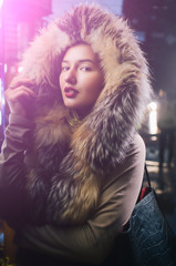 Beautiful caucasian girl  in winter clothes on the street, attractive teenager portrait with a fur jacket , she's watching a timeline of transport on  an electronic screen illuminated by a violet