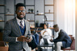© alfa27 - Black businessman wearing grey tuxedo holding smartphone during work time in modern office, business partners meeting background. Look at camera and smile. Business people concept