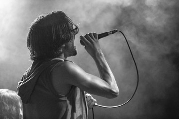 A male musician is viewed from the side as he sings, with open mouth in microphone and interacts with the audience, black and white with copy space