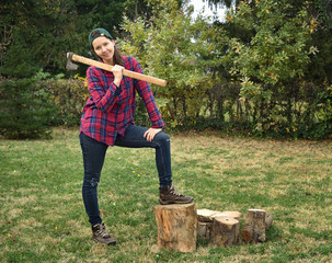 Smiling female lumberjack holding a huge axe on her shoulder standing with one leg on the log