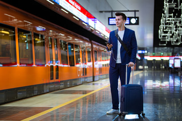 Young stylish handsome man in suit with suitcase standing on metro station holding smart phone in hand, scrolling and texting, smiling and laughing. Futuristic bright subway station. Finland