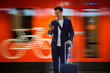 © Suzi Media  - Young stylish handsome man in suit with suitcase standing on metro station holding smart phone in hand, scrolling and texting, smiling and laughing.  Orange train passing by. Futuristic bright subway