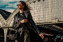 Outdoor Fashion Portrait Of Young Confident Model, Woman Wearing Trendy Black Leather Trench Coat, Dress, Glasses, Wrist Watch, With Small Hobo Bag, Walking In Street Of City. Copy, Empty Space