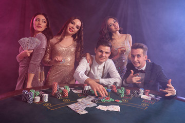 Friends playing poker at casino, at table with stacks of chips, money, cards on it. Celebrating win, smiling. Black, smoke background. Close-up.