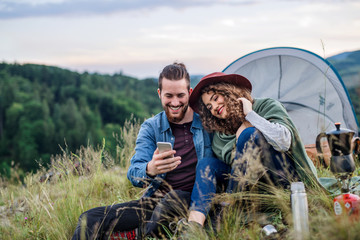 Wall Mural - young tourist couple travellers with tent shelter sitting in nature, using smartphone.