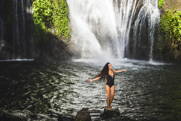 Young slim brunette woman with curly hair enjoying in lagoon of huge tropical waterfall Banyumala in Bali. Wearing in black swimsuit. Happy vacations in Indonesia. Wanderlust travel concept.