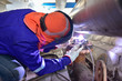 Worker in blue uniform,leather gloves,welding mask. He is sitting and welding the workpiece with a Tungsten Inert Gas Welding process (TIG).