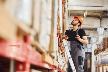 Storage worker stands on the ladder in uniform and notepad in hands and checks production