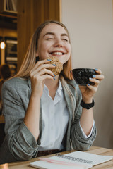 Lifestyle Portrait of smiling young casual blonde woman eating a cookie and drinking coffee, tea in cafe. Happy girl eating a dietetic gluten free cookie