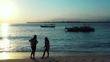 Two Girls Waiting On Sandy Beach For Sunset Moments, To Watch Reflecting Of Last Rays Over Calm Lagoon Where Boats Are Anchored In Thailand