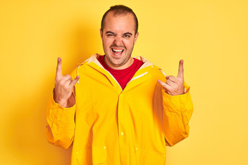 Sticker - Young man wearing rain coat standing over isolated yellow background shouting with crazy expression doing rock symbol with hands up. Music star. Heavy concept.