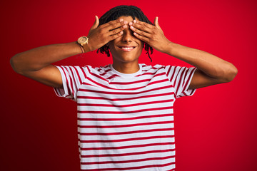 Wall Mural - Young afro man with dreadlocks wearing striped t-shirt standing over isolated red background covering eyes with hands smiling cheerful and funny. Blind concept.