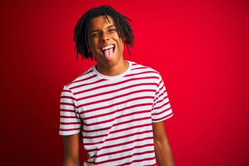 Wall Mural - Young afro man with dreadlocks wearing striped t-shirt standing over isolated red background sticking tongue out happy with funny expression. Emotion concept.